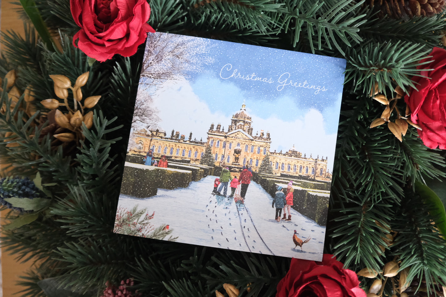 Christmas card with a snowy scene of Castle Haword, Yorkshire, including people and the building, surrounded by festive greenery and red roses.