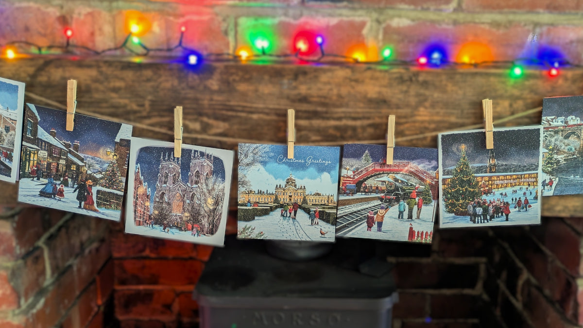 A string of small square printed cards clipped with wooden clothespins hangs across a rustic wooden mantel in a brick fireplace. Multicolored holiday string lights glow above, and the cards show winter city and festive scenes.